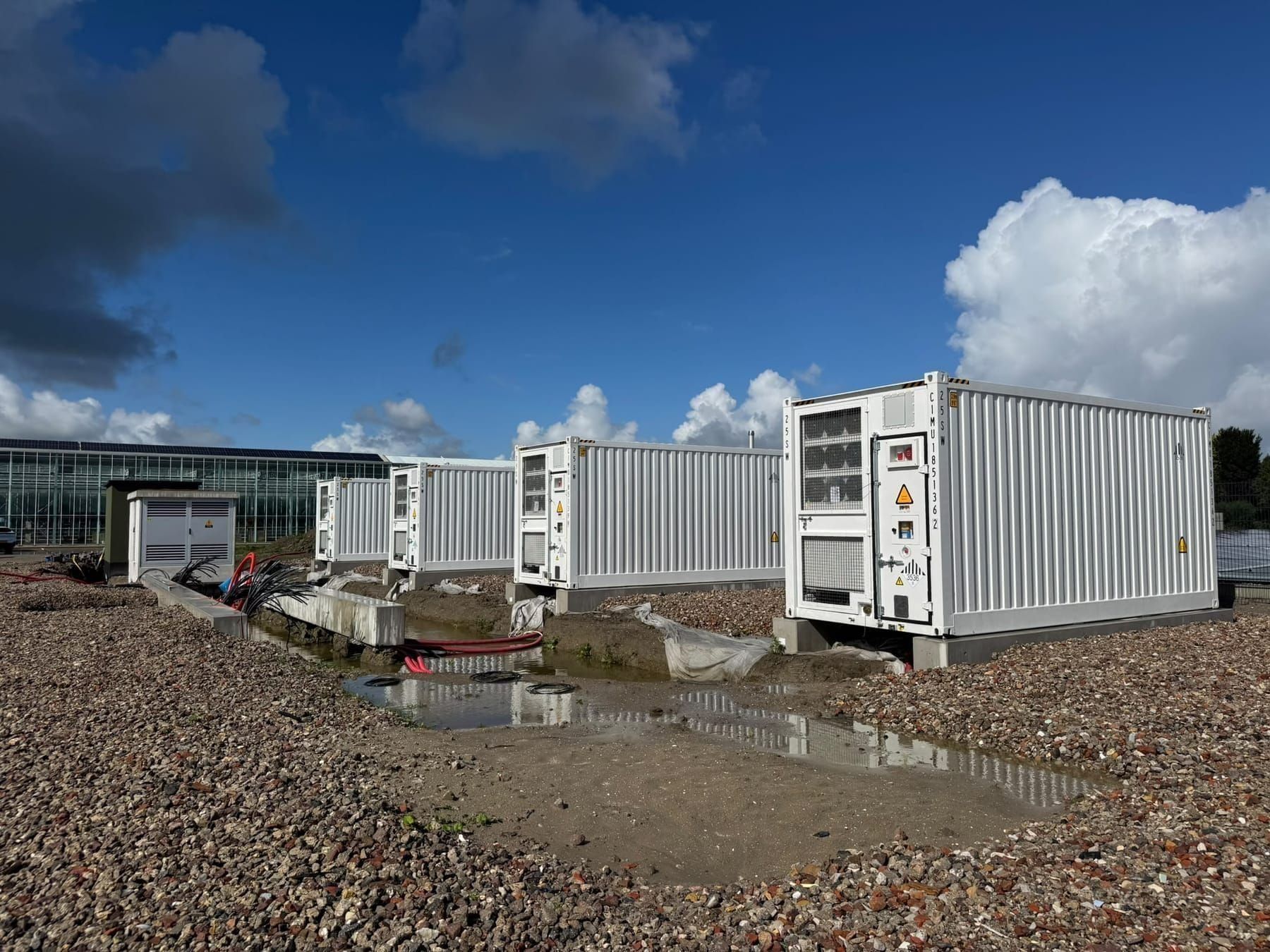 Rij batterijcontainers op hun fundering, met de kassen op de achtergrond